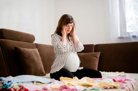 Pregnant woman worries above baby clothes at home on sofa